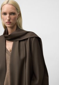 Blonde Frau, die einen braunen oversized Blazer über den Schultern trägt, einen beigen V-Ausschnitt-Pullover und eine goldene Anhänger-Halskette vor einem schlichten Hintergrund.