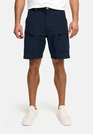 Mann trägt marineblaue Cargo-Shorts mit mehreren Taschen, weißen Gürtel, weiße Turnschuhe, steht vor einem schlichten Hintergrund.