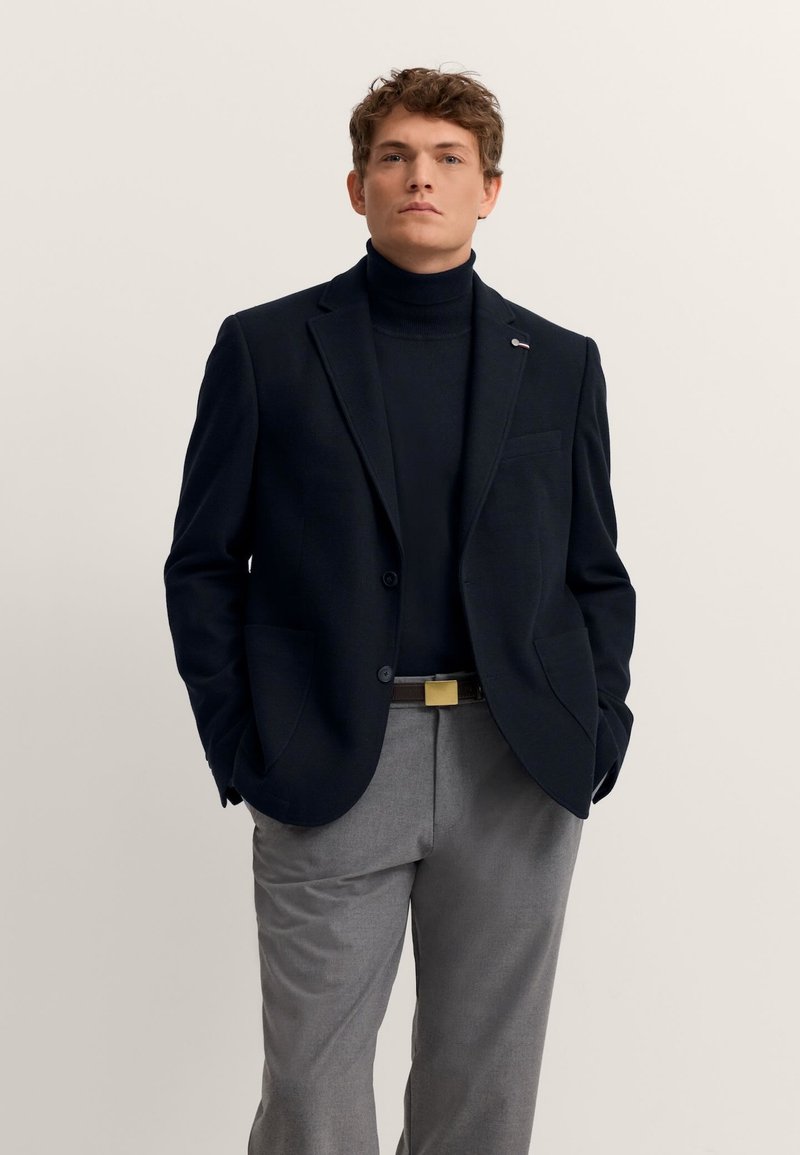 Marineblazer mit strukturierter Oberfläche, Ein-Knopf-Verschluss und zwei Seitentaschen, getragen über einem schwarzen Rollkragenpullover und grauen Hosen, mit einer goldenen Gürtelschnalle.