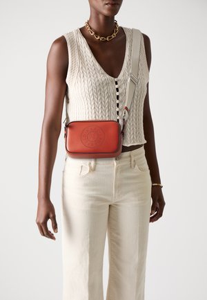 Rote Leder-Crossbody-Tasche mit gesteppten Designdetails, silberner Hardware, verstellbarem Tragegurt und rechteckiger Form. Getragen über einem cremefarbenen Sweater und hellen Jeans.