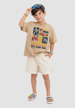 Garçon portant un t-shirt beige avec des icônes sur le thème de la mer, un short blanc rayé, des bracelets et un collier de perles colorées, tenant des lunettes de soleil bleues.