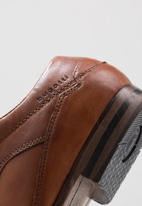 Chaussure en cuir marron avec une texture lisse, des détails de couture subtils, une semelle en caoutchouc noire et le nom de la marque "bugatti" embossé sur le côté.