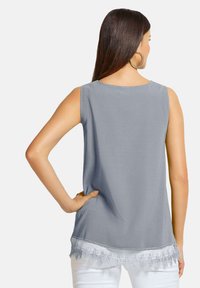 Graues ärmelloses Oberteil mit rundem Halsausschnitt, das am Saum mit einer Spitzenverzierung versehen ist und eine glatte Textur aufweist. Kombination mit weißen Hosen.