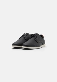 Zapatos negros de cordones con un upper texturizado, suela crema y acentos marrones en contraste cerca del talón. Diseño clásico con punteras redondeadas.