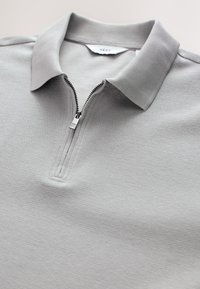 Graues Polohemd mit Reißverschluss aus strukturiertem Stoff, mit klassischem Kragen und silbernem Reißverschluss. Etikett innen am Kragen sichtbar.