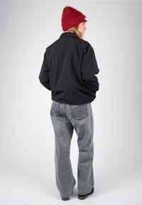 Femme portant un bonnet rouge, une veste noire et un jean gris ample, debout légèrement tournée sur le côté sur un fond uni.