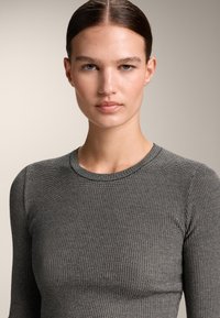 Persoană tânără cu părul dat pe spate, purtând un top gri texturat, cu mâneci lungi, bine ajustat, privind drept înainte cu o expresie neutră.