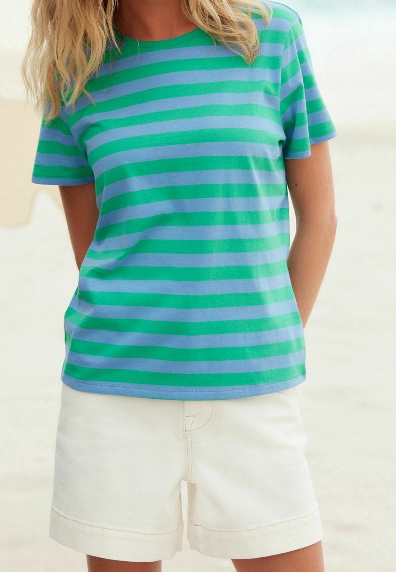 Femme aux cheveux blonds portant un t-shirt rayé bleu et vert et un short écru, debout avec les mains dans le dos.