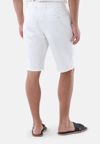 Weißes Baumwollshorts mit taillierter Passform, ausgestattet mit Gesäßtaschen und dezenten Stichdetails, kombiniert mit dunklen Slides. Einfaches und funktionales Design.