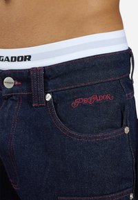 Denim jeans featuring dark blue fabric with contrasting red stitching, embroidered "PEGADOR" logo, and a white elastic waistband with black lettering.