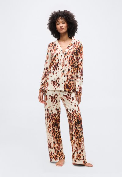 Γυναίκα που στέκεται ξυπόλυτη φορώντας μακριά μπεζ πουκάμισα με κουμπιά και παντελόνι με καφέ και μαύρο μοτίβο λεοπάρ.