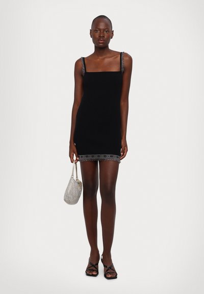 Robe mini noire à côtes avec décolleté carré, bordure en dentelle argentée et bretelles délicates. Le mannequin tient un sac à main en maille argentée.
