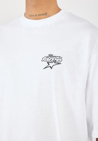 Camiseta de algodón blanca con un estampado gráfico negro que dice "el placer de la fama" con un acento de estrella, cuello redondo y mangas cortas.