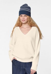 Ecrufarbener, strukturierter V-Ausschnitt-Pullover mit lässiger Passform, kombiniert mit einer navyblauen Strickmütze mit einem weißen geometrischen Muster.