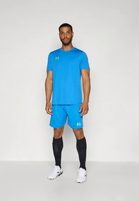 Vyras stovi vilkėdamas ryškiai mėlyną sportinę aprangą, juodas iki kelių kojines ir baltus sportbačius prieš paprastą šviesiai pilką fono.