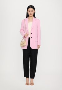 Svijetlo ružičasti krojeni blazer s crnim gumbima, kremasti top, crne hlače i mala bež torba. Model nosi gojzerice u boji kože. 