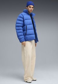 Ung mand iført en blå pufferjakke, matchende blå hue, brede cremefarvede bukser og hvide sneakers, stående imod en grå baggrund.