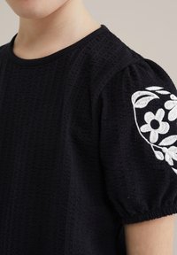 Crna teksturirana košulja s kratkim napuhanim rukavima, ukrašena bijelim cvjetnim vezom na jednom rukavu i okruglim izrezom.