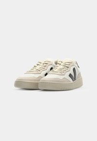 Beige and white sneakers with a textured finish, featuring black logo accents, perforated sections, and a thick rubber sole.