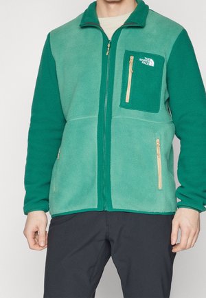 Zielona kurtka z polaru z kieszenią na piersi na zamek, złotymi elementami i kontrastującymi teksturami, z kołnierzem stójką i zapięciem na zamek.