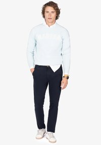 Hombre con sudadera azul clara sobre camisa blanca, pantalones azul marino oscuros, zapatillas blancas, reloj de pulsera, de pie con las manos en los bolsillos.