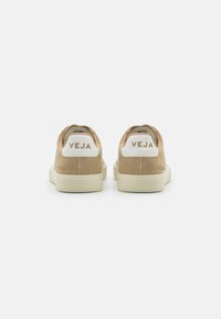 Beige suede low-top sneakers with white heel tab and embossed "VEJA" logo. Soft texture and contrasting rubber sole.