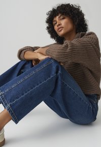 Maglione a coste marrone con maniche lunghe, abbinato a jeans di denim blu larghi. Modella seduta, con scarpe chiare e capelli ricci.