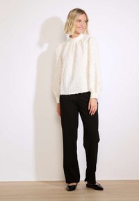 Blusa bianca a maniche lunghe con texture e scollatura arricciata, abbinata a pantaloni neri a gamba dritta e ballerine nere su uno sfondo chiaro.