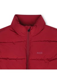 Veste rembourrée rouge avec un col montant, dotée d'une fermeture éclair à l'avant et de coutures horizontales. Le logo est subtilement imprimé sur la poitrine gauche.