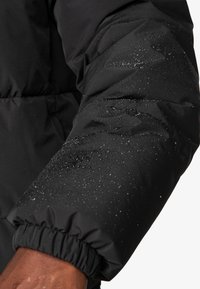 Sort pufferjakkeærme med elastisk manchett, prydet med vanddråber på en glat, matt stofoverflade, der fremviser et quiltet design.