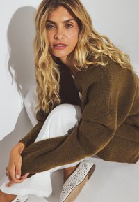 Suéter marrón texturizado con puños acanalados, usado sobre pantalones de lino blancos. Presenta intrincados zapatos planos de tejido blanco. Maquillaje natural y cabello despeinado.