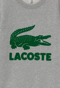 Szara bluza z dużym zielonym wizerunkiem krokodyla oraz napisem "LACOSTE" w pogrubionych zielonych literach poniżej. Standardowy krój z okrągłym dekoltem.