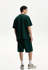 Prevelik zeleni bombažni majica in ujemajoče se kratke hlače, ki imajo sproščen kroj, kratke rokave in vidne šive.