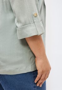 Μανίκι πουκάμισου ανοιχτού πράσινου με υφή, διπλωμένο προς τα πάνω και με μπεζ κουμπί, ακουμπισμένο πάνω σε λυγισμένο χέρι και μπλε τζιν.