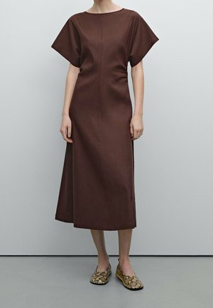 Woman wearing a brown midi dress with short wide sleeves paired with snake-print flat shoes, standing against a plain gray wall.