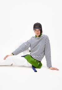 Maglia a maniche lunghe a righe con righe blu navy e bianche sopra un colletto verde, abbinata a pantaloni bianchi e a un cappellino nero con la scritta "PERMANENT".