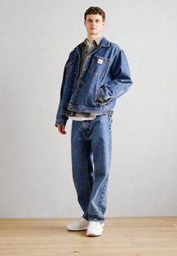 Veste en jean bleu clair, avec un patch sur la poche poitrine, associée à un jean bleu clair à coupe décontractée et des baskets blanches.