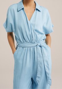 Ljusblå, kortärmad jumpsuit i ett mjukt tyg, med en invecklad design, bälte i midjan och sidofickor. Slät textur och avslappnad passform.