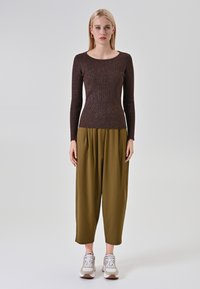 Femme se tenant contre un fond uni, portant un pull à manches longues marron, un pantalon large olive et des baskets blanches.