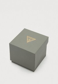 Caixa quadrada cinzenta com um logótipo triangular dourado apresentando a palavra "GUESS" e um ponto de interrogação no centro superior.