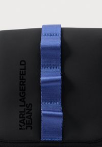 Μαύρη τσάντα με λεία υφή και λαβή από κορδέλα σε αντίθετο μπλε χρώμα. Διαθέτει ανάγλυφη επιγραφή "KARL LAGERFELD JEANS" στο πλάι.