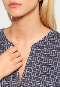 Dunkelblaue, gemusterte Bluse mit Knopf-Front-Design, hexagonalen Mustern und einer weichen Textur, die angenehm auf der Schulter eines Modells ruht.