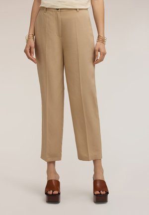 Mujer con pantalones beis de talle alto y pierna recta, combinados con sandalias marrones de plataforma y puntera abierta, y pulseras rígidas doradas.