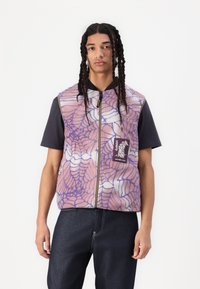 Fleece vest with a purple and pink spiderweb pattern, zip closure, and a logo patch. Worn over a dark t-shirt and paired with dark jeans.
