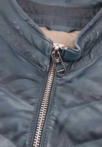 Lederjacke in dunklem Petrol mit glatter Textur. Verfügt über einen silbernen Metallreißverschluss und einen Stehkragen, der mit gesteppten Nähten verziert ist.