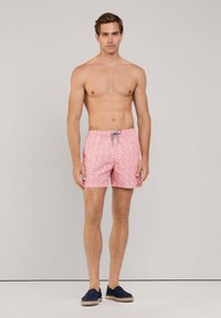 Costumi da bagno rosa con un motivo geomentrico. Realizzati in tessuto leggero, presentano una vita elastica con cordoncino. Il modello posa a piedi nudi.