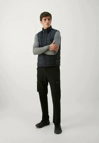 Mornarsko podstavljena prsluk s visokim ovratnikom, zatvoren na zip, nošen preko sivoj košulje dugih rukava, u kombinaciji s crnim cargo hlačama i crnim cipelama.