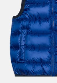 Nieprzemakalna niebieska kamizelka z pikowaną teksturą, wyposażona w zamek błyskawiczny z przodu i boczną kieszeń. Krawędzie wzmocnione ciemniejszym materiałem.