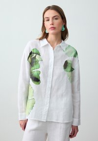 Camicia bianca in lino caratterizzata da grandi stampe floreali verdi sul lato sinistro, con colletto classico e chiusura con bottoni.