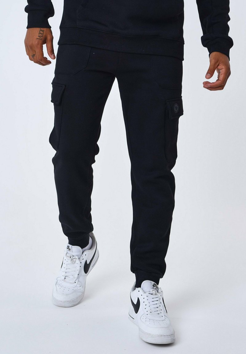 Pantalones de chándal negros con perneras ajustadas, bolsillos tipo cargo a los lados y puños acanalados. Combinados con zapatillas blancas con detalles en negro.